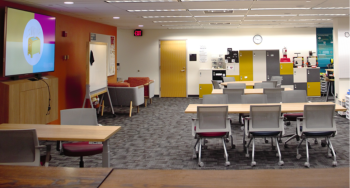 Large open space with multiple tables, chairs, and a monitor with lockers on the back wall.