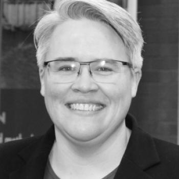 A smiling white person with short, straight grey hair and glasses. I'm wearing a red crewneck shirt under a black blazer jacket, and have a small bronze-ish lapel pin.