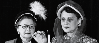 A black and white photograph of two women in front of a long tapered candle, one woman in the act of lighting the candle.