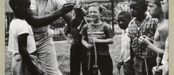 A black and white photograph of a man showing a group of boys from a community center recreation program how to use a bow and arrow