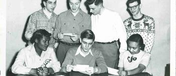 A group of seven students gather to look at pieces of paper. Three students are seated, with four students standing behind them.