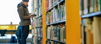 A young man wearing a winter coat and hat browses the books in the brightly lit Architecture Library. 