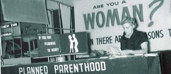 A black and white photograph shows the Planned Parenthood information booth at the Minnesota State Fair circa 1955-1960. The sign on the booth reads Are you are woman? Planned Parenthood Can Help You. A woman volunteer sits behind the table. 