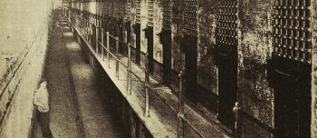 A black and white photo of prison cellblocks at Sing Sing prison in New York. A man stands to the side.
