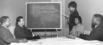 A group of social workers seated around a table at a training course. A woman is standing and using a pointer to show words on a blackboard.