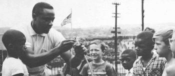 A youth worker shows a group of boys how to string a bow. The boys are holding bows and appear to be watching the demonstration intently. The group is standing outside with a view of a city, the American flag, and utility poles in the background.