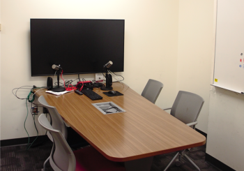 Small private room with table, monitor, podcasting equipment such as microphones, and four chairs.